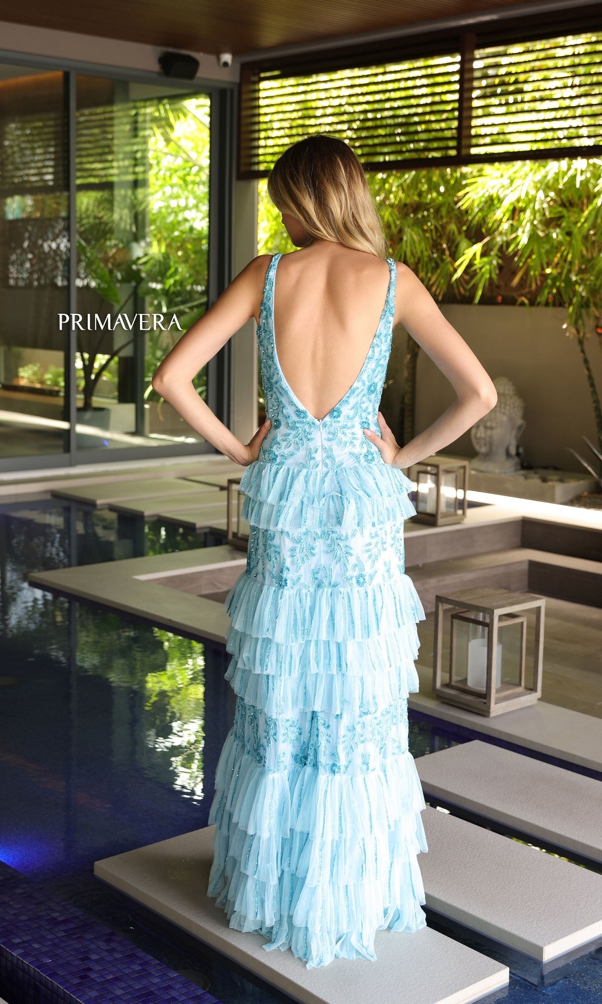 A woman stands by an indoor pool, facing away from the camera, wearing the Primavera Beaded Long Ruffled Prom Dress 4106—a light blue backless gown with tiered ruffles and her hands on her hips.